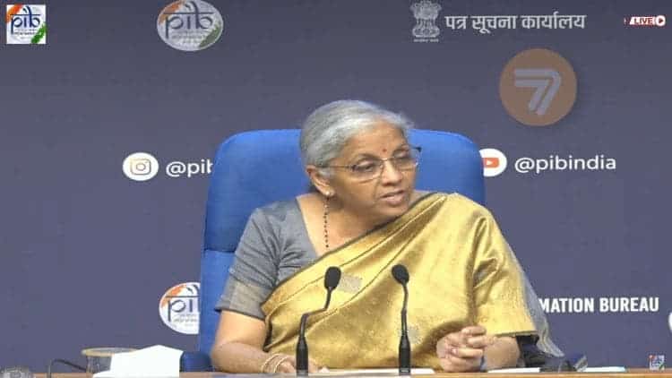 Smt. Nirmala Sitharaman sitting on a chair and speaking on two microphones positioned on the table in front of her, against a background provided by Press Information Bureau India.