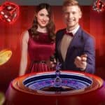 Two game hosts stand beside a roulette wheel. Dice and golden chips fly around them.