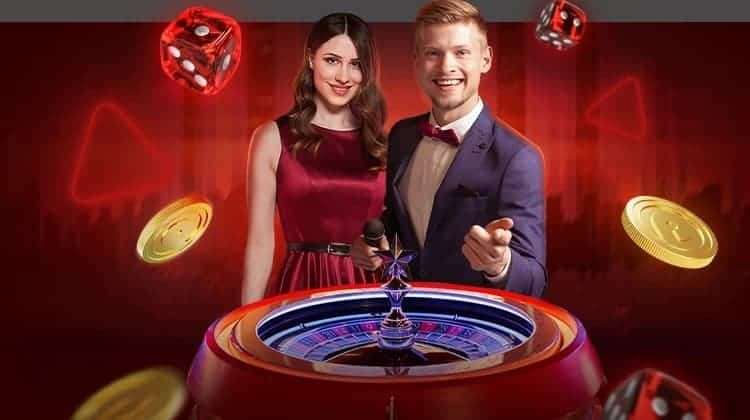 Two game hosts stand beside a roulette wheel. Dice and golden chips fly around them.